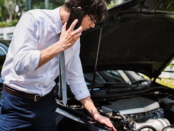 Ein junger Businessmann starrt auf den Motor des Autos und ruft gleichzeitig mit seinem Smartphone an. 